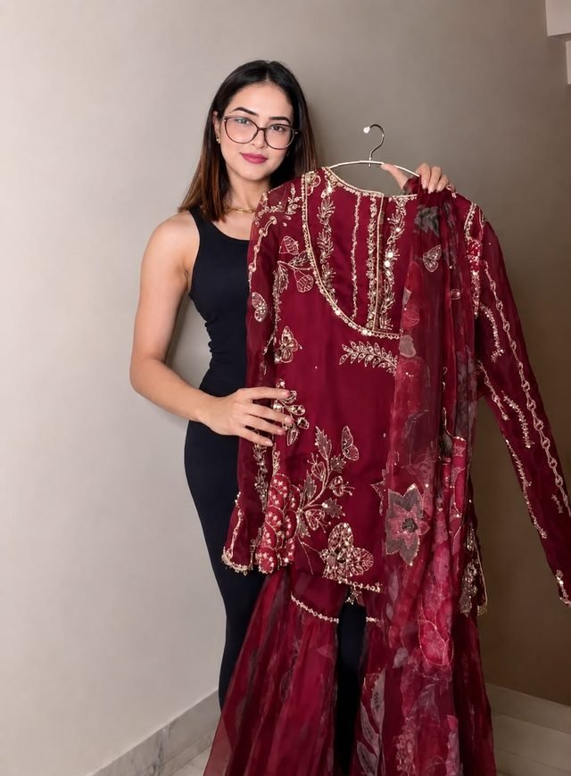 Woman in glasses holding a maroon embroidered sharara set with a sheer printed dupatta on a hanger