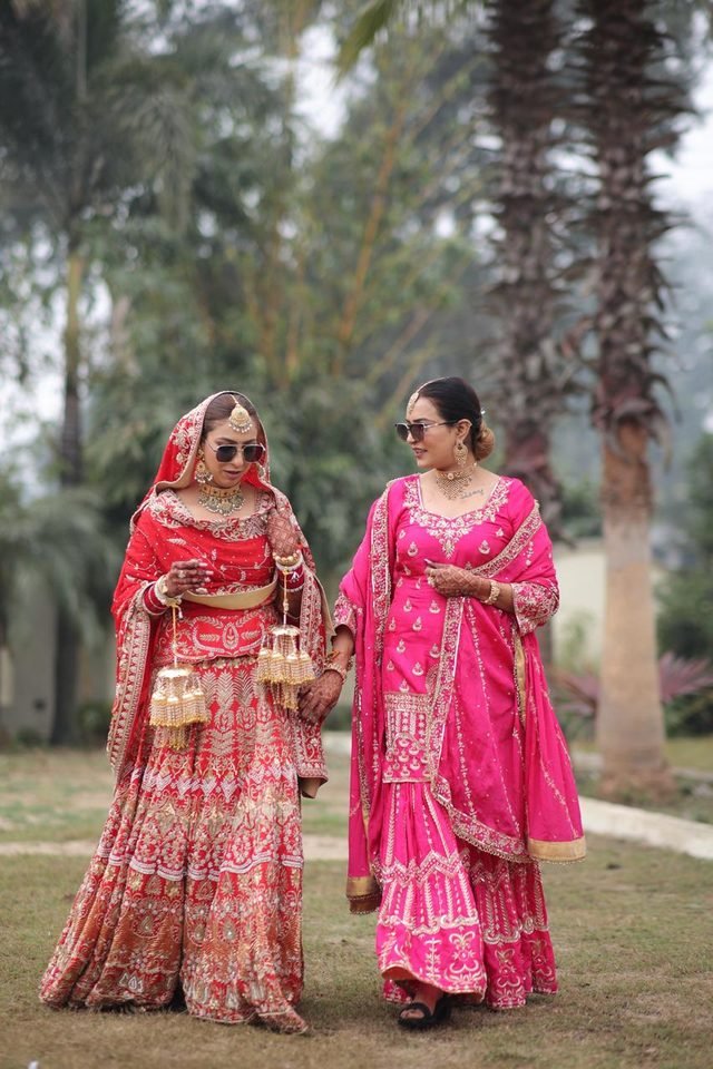 Two women walking outdoors, one in a red embroidered bridal lehenga and one in a pink embroidered sharara set